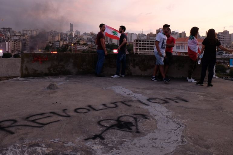 Young men and women enjoy the view atop an abandoned theater in downtown Beirut as smoke rises in the background during large street protests