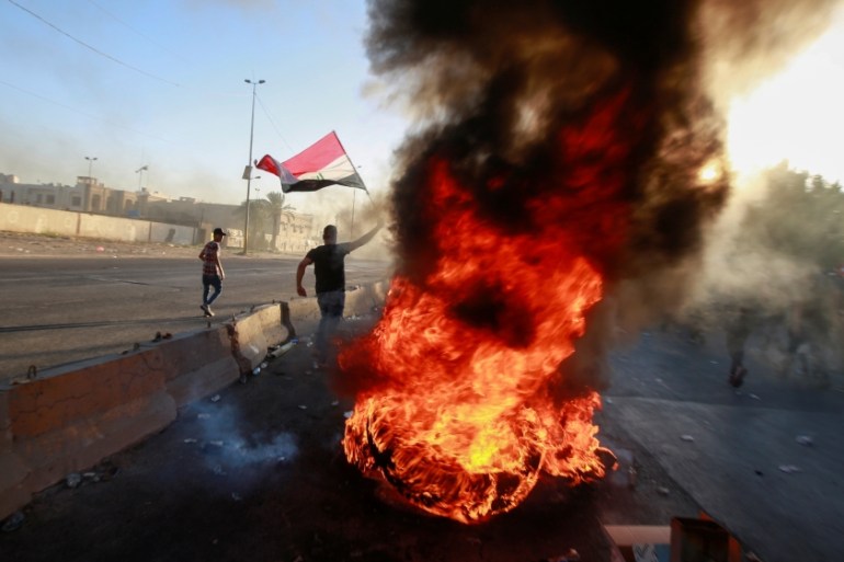A demonstrator holds the Iraqi flag near burning objects at a protest during a curfew, three days after the nationwide anti-government protests turned violent, in Baghdad