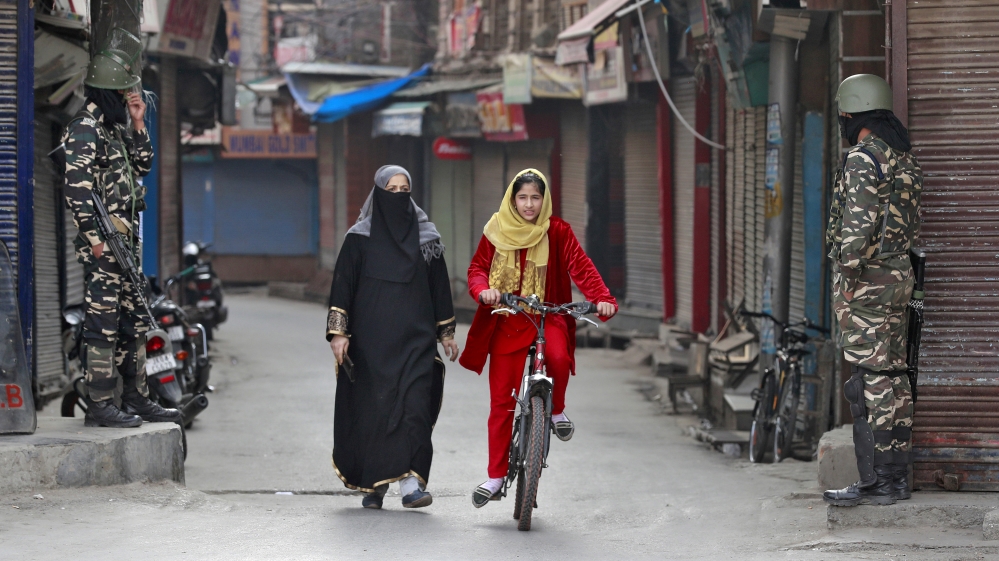 A Kashmir girl rides her bike past Indian security force personnel standing guard in front closed shops in a street in Srinagar