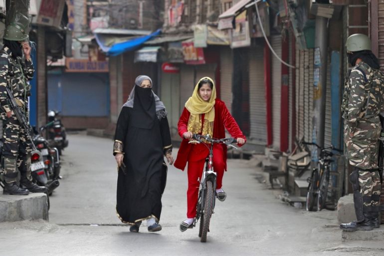 A Kashmir girl rides her bike past Indian security force personnel standing guard in front closed shops in a street in Srinagar