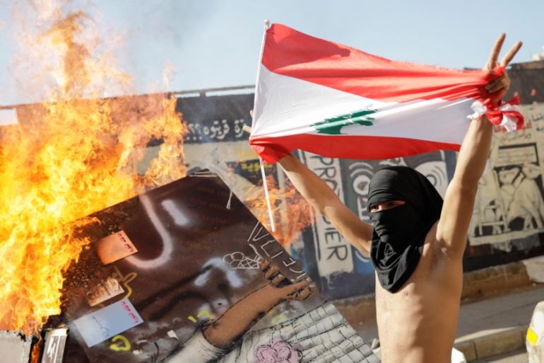 A masked protester holding a Lebanese flag walks past a burning barricade during ongoing anti-government protests in Beirut