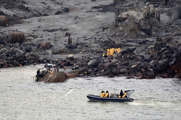 Rescuers on the White Island volcano on a mission to retrieve victims of the eruption. The land is grey and dark. There is a second group of rescuers in a dinghy on the water.