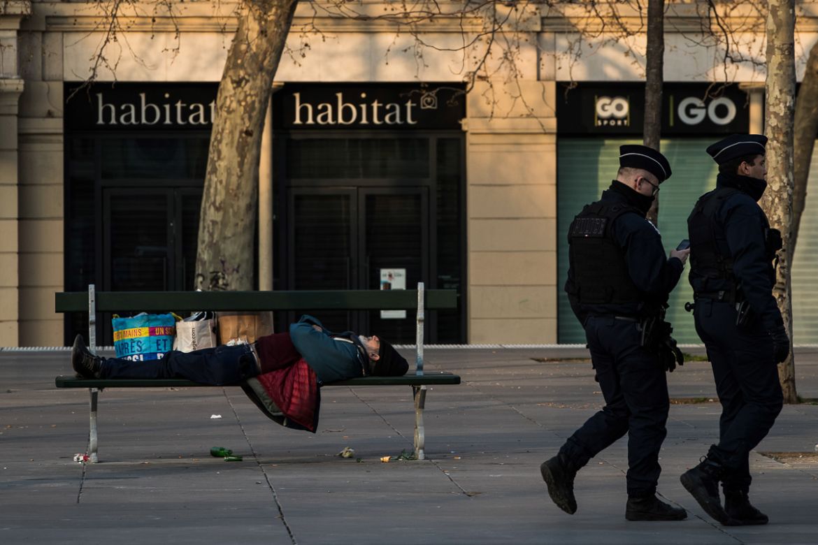 Policemen walks by a homeless man sleeping on a bench on March 17, 2020 in Paris, as a strict lockdown comes into effect in France to stop the spread of COVID-19, caused by the novel coronavirus. - A