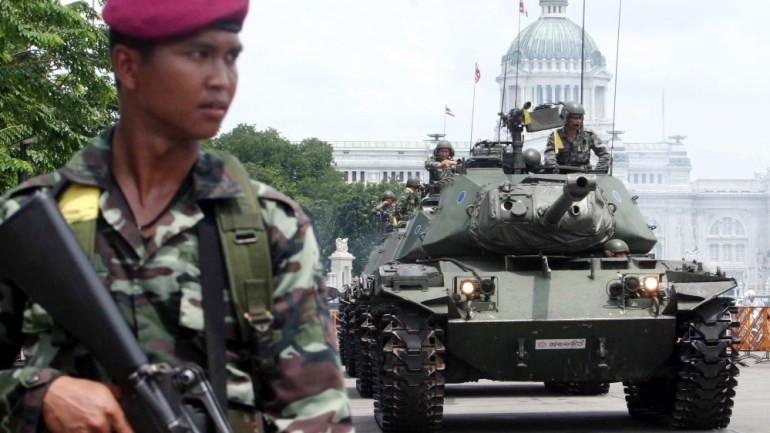 Thailand coup. A soldier with a beret is in the foreground wearing a red beret and holding a weapon. He is slightly out of focus. Behind him and clearly in focus is a large tank with a soldier half-sitting inside it. Further off in the background are other military personnel and equipment as well as the Dusit Palace.