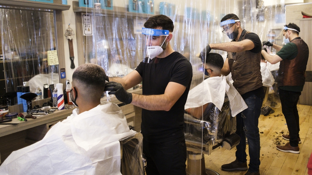 Barbers wear protective equipment and clients sit in chairs separated by plastic dividers at Hell’s Kitchen Barbers on the first day that hair salons could reopen as part of Phase 2 in New York, New Y