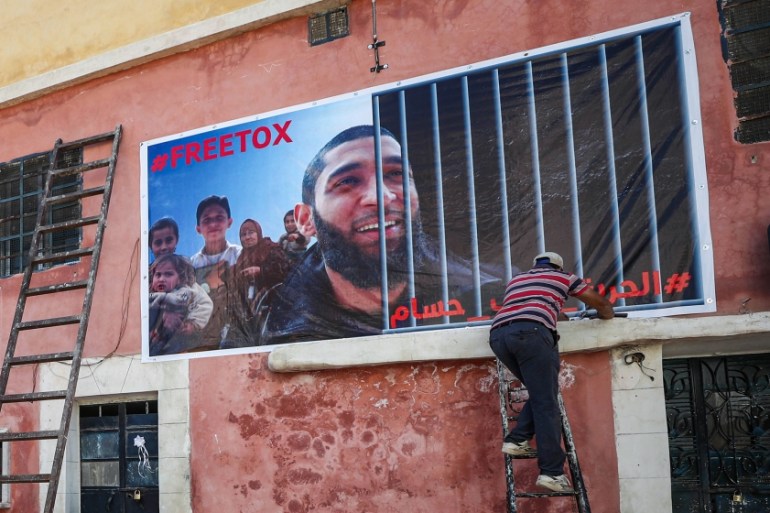Men fix up a banner on the side of a building calling for the release of Tauqir Sharif, a self-described aid worker stripped of his British nationality and detained by Hayat Tahrir al-Sham (HTS) jihad