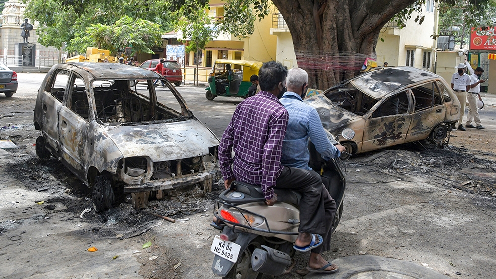 Motorists ride past burnt vehicles in Bangalore on August 12, 2020, after violence broke out overnight in Devara Jevana Halli area following a 