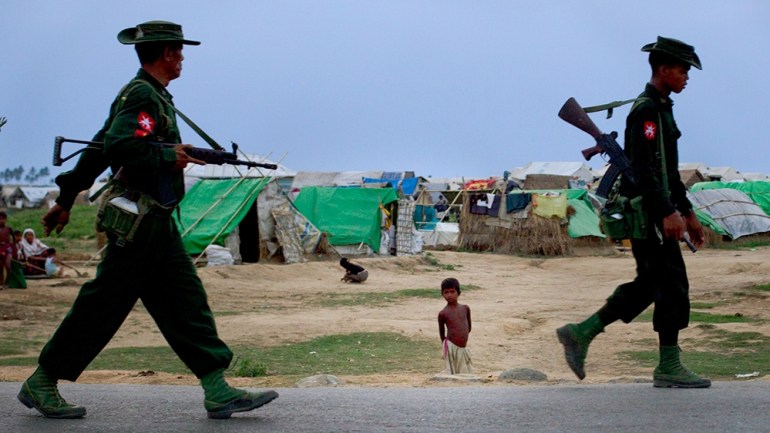 In this May 13'' 2013 photo, an internally displaced Rohingya boy, center watches army soldiers on foot-patrol in the foreground of makeshift tent camps for Rohingya people in Sittwe, northwestern Rakh