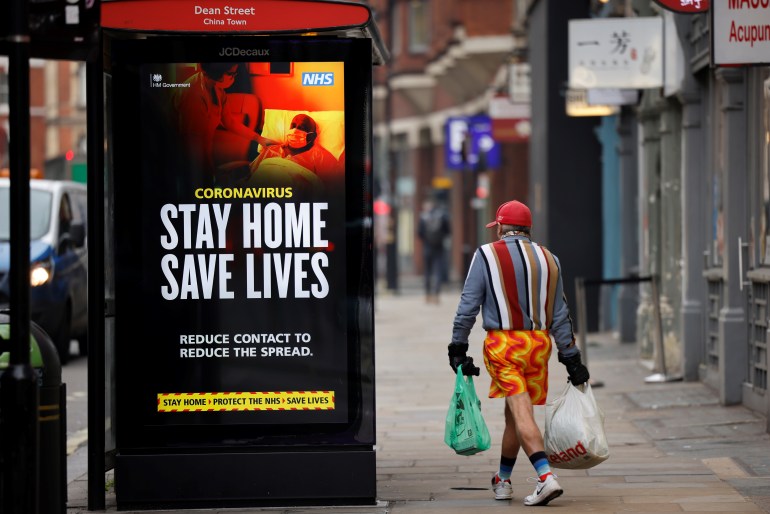 A shopper walks past NHS signage promoting "Stay Home, Save Lives" on a bus shelter