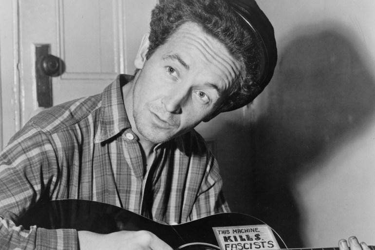 Woody Guthrie is seated playing a guitar that has a sticker attached reading: This Machine Kills Fascists [Library of Congress]