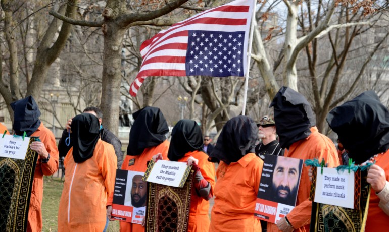 Activists in orange prison jumpsuits protest with signs next to an American flag