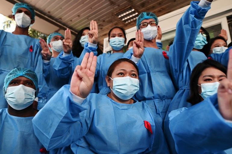 Medical workers in blue medical gowns and face masks take part in mass protests against Myanmar's military. The photo shows a group of workers, a woman and a man, holding up a three-finger salute.