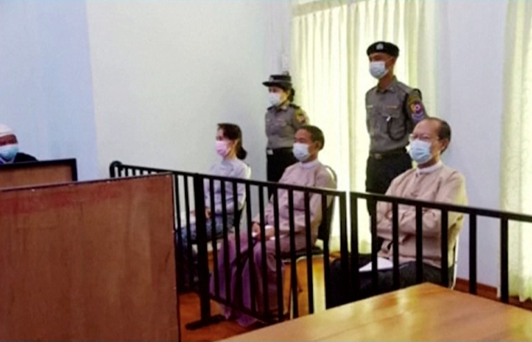 Aung San Suu Kyi siège au tribunal avec une policière derrière elle lors de sa première apparition publique depuis le coup d'État du 1er février 2021.
