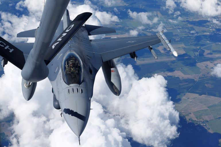 Um F-16 dos EUA se preparando para um reabastecimento em pleno voo.  As nuvens e a terra são visíveis abaixo do avião.