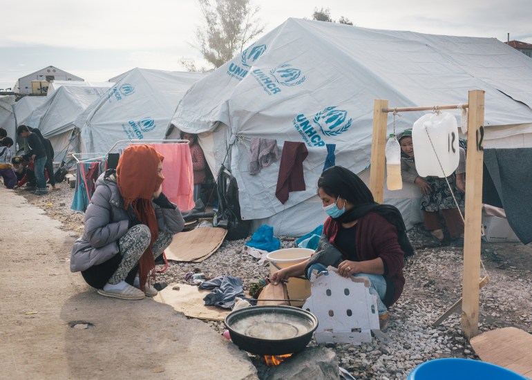 Two woman crouch and cook in front of an open fire near tents in a refugee camp