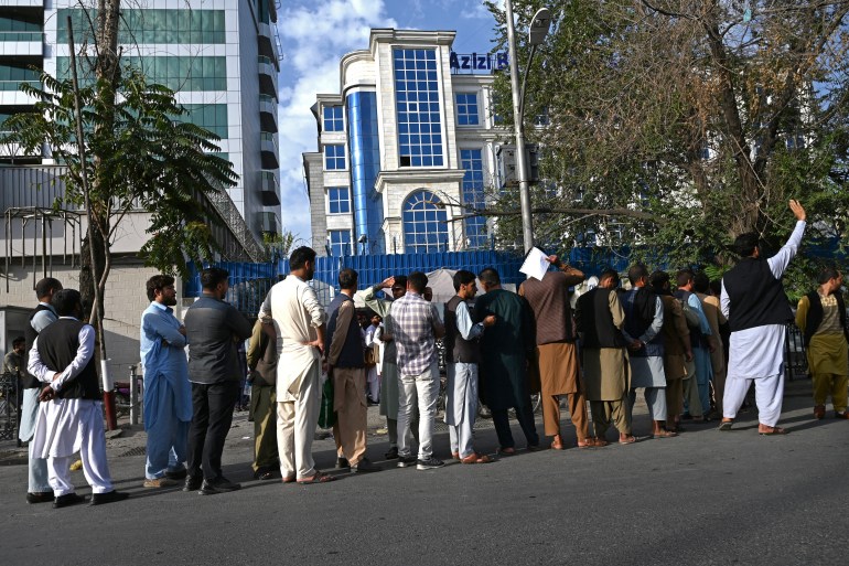 Afghan people stand in a queue as they wait for their turn to collect money from an ATM in front of a bank along a roadside in Kabul, Afghanistan
