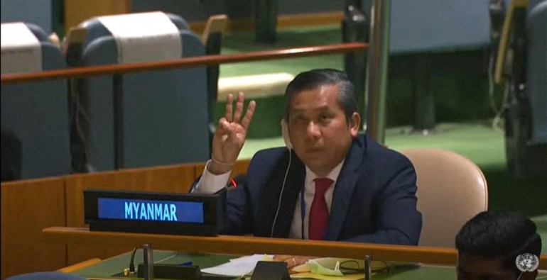 O diplomata Moe Kyaw Tun fazendo a saudação de três dedos do movimento anti-golpe de Mianmar em uma reunião da ONU cinco dias depois que os militares tomaram o poder.  Ele está sentado na mesa de Mianmar na Assembleia Geral da ONU