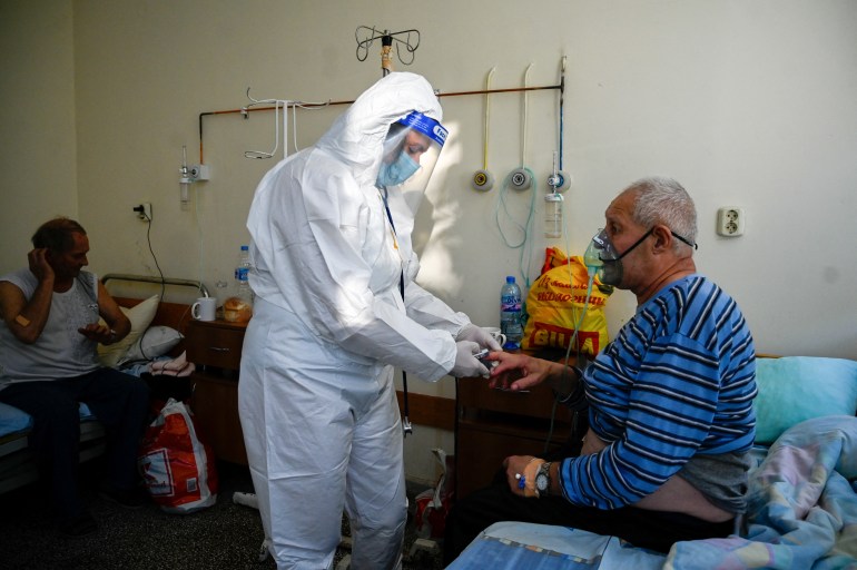 A doctor in a white full body suit checks on a patient wearing a respiration mask in a hospital.