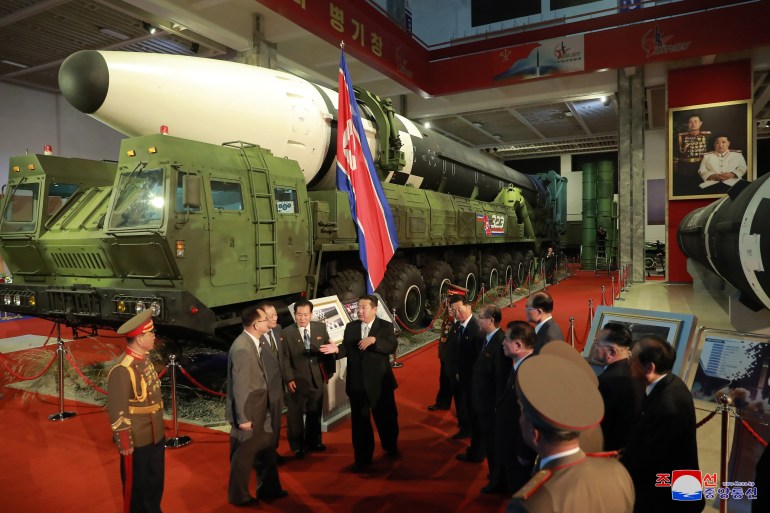 North Korean leader Kim Jong Un speaking to officials at a defence exhibition in October 2020 in front of a rocker with a white nose cone on a launcher, and a North Korean flag