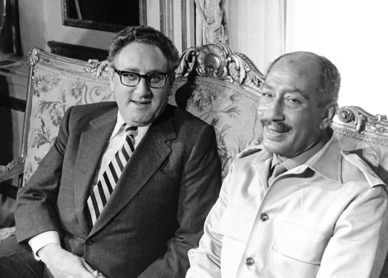 Henry Kissinger and Anwar Sadat seated on a floral couch.