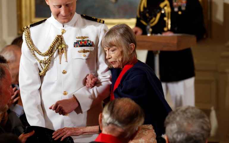 Joan Didion holds the arm of a man in military dress while being escorted to her seat.