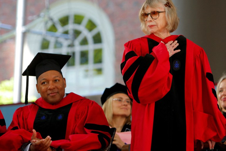 Wynton Marsalis, seated in cap and gown, applauds Joan Didion, robed and standing, on a stage.
