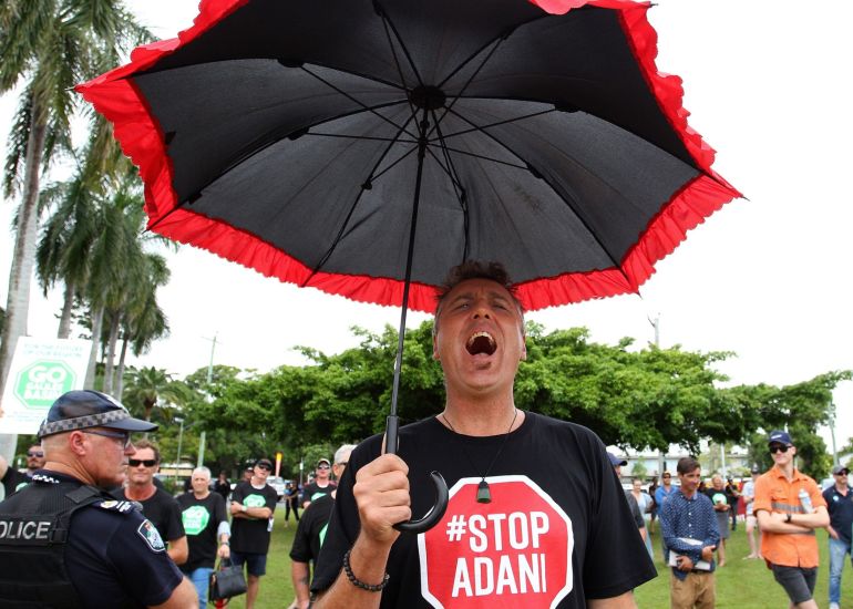 An activist at a rally in Mackay, Queensland, Australia, wears a black T-shirt with "STOP ADANI" written on it in white letters in a red STOP sign.  He holds a black umbrella with red folds around it.