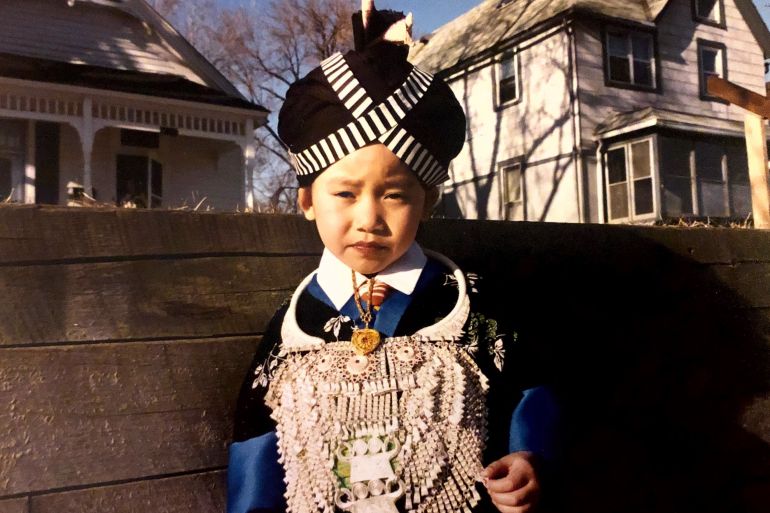 A young girl wearing a traditional Hmong dress stands in front of a house in America