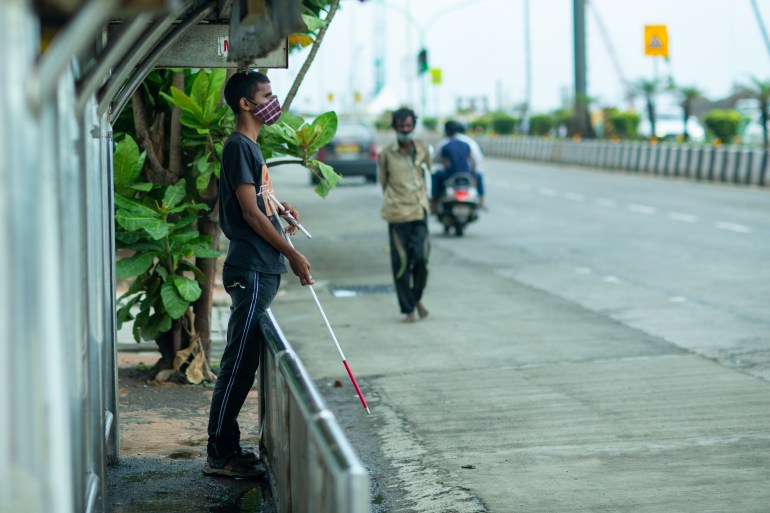A visually impaired young man holding a white cane leans against a bus stop barrier at the edge of the pavement.