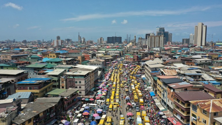 An aerial photograph of a congested road in Lagos. The middle of the road is crowded with yellow buses and the sides of it with people.