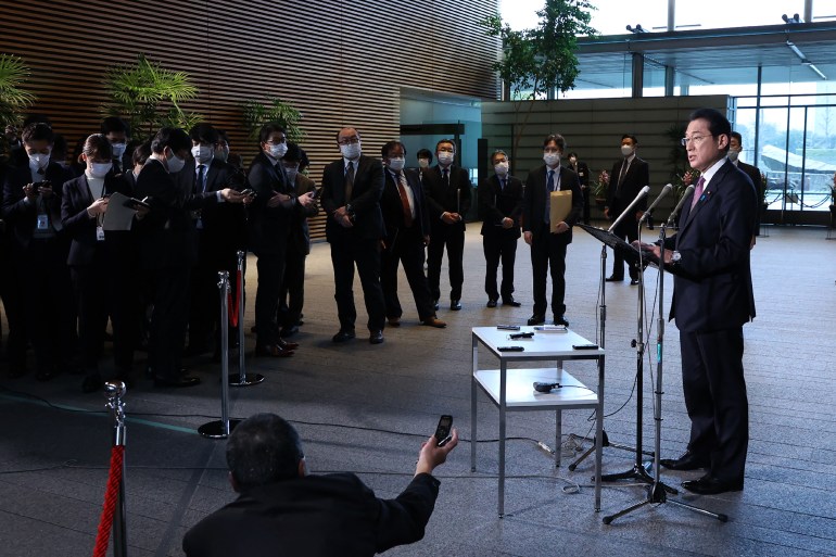 Japan's prime minister stands behind a lectern on the right  answering  questions from  journalists  gathered  some distance from him in a semi-circle