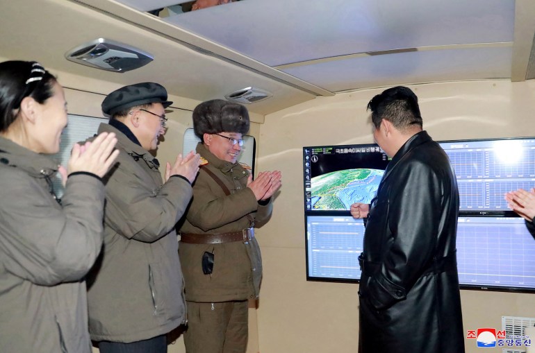 Kim Jong Un on the right in a long black leather jacket discusses a rocket launch with official including his sister Kim Yo Jong who stands on the right and claps enthusiastically