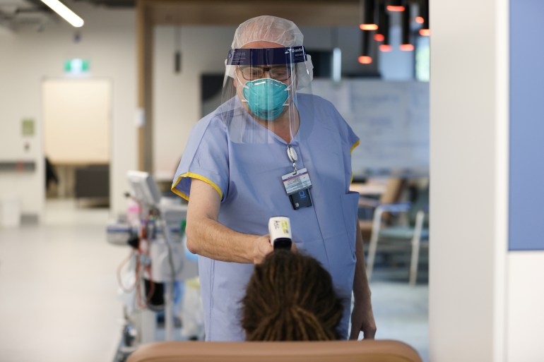 A nurse in an N95 mask and clear visa stands in front of a patient to take their temperature in a Covid clinic