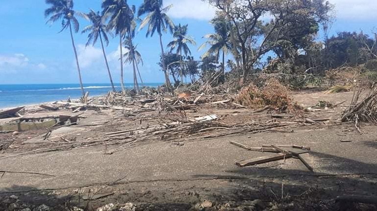 A beach in Nuku'alofa, with palm trees behind and a blue sky, covered with volcanic ash and debris from the tsunami