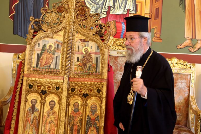 Archbishop Chrysostomos II stands next to a pair of ornate, gilded doors that guard the altar of a church