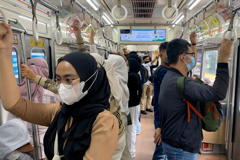 A masked woman in a black headscarf and peach-colored jacket stands with other passengers on a Jakarta metro train while COVID cases decrease to several hundred a day