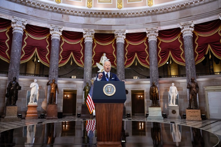 President Joe Biden is seen speaking on a podium with red drapes, statues and marble columns in the background.