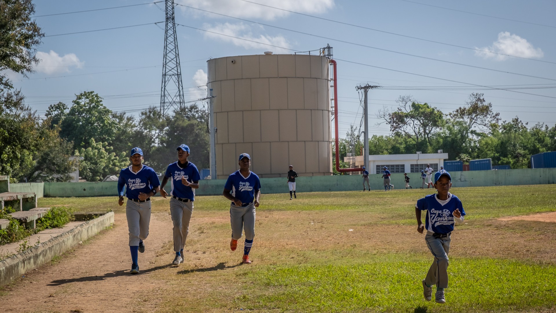 Boys playing baseball jog on a pitch that lies beside chemical storage containers
