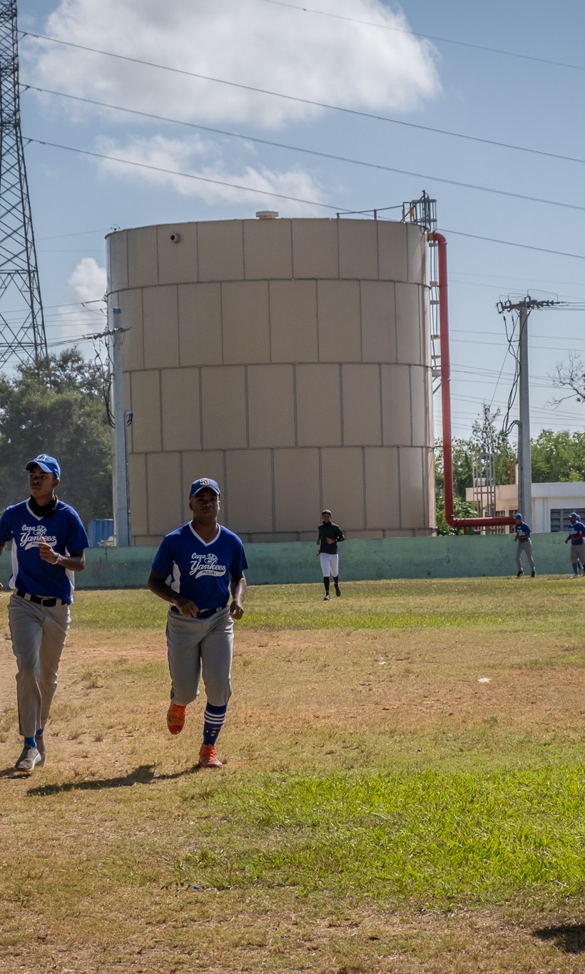Boys playing baseball jog on a pitch that lies beside chemical storage containers