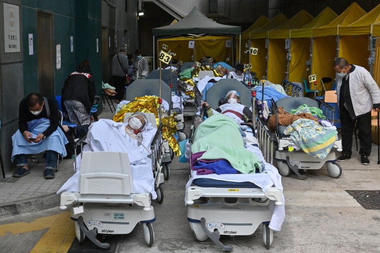 Patients lie beneath blankets on hospital beds outside a Hong Kong medical facility with some sitting on chairs, blankets around their shoulders.