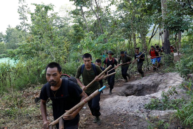 A group of men with rudimentary wooden weapons queue through a forest meadow while training from the Myanmar People's Defense Forces (PDF)