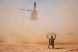 A French solider, on one knee, guides in a helicopter in Mali as dust swirls and obscures the sky