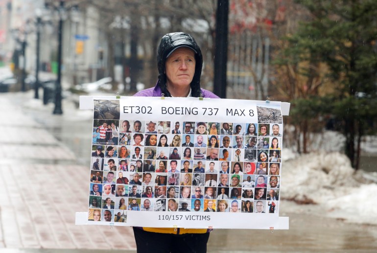 Moore wearing a helmet is standing in the street, holding a poster showing the faces of the victims of the crash