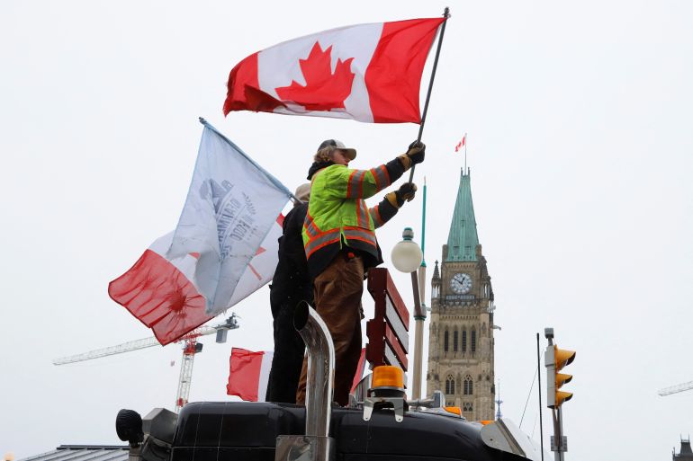 A man stands on the top of a truck wearing a high vis jacket and flying a Canadian flag during the truckers' protest