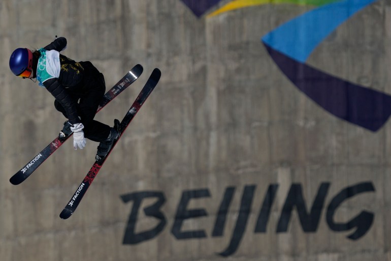 Eileen Gu pictured mid-air in front of a gray concrete wall emblazoned with the Beijing Winter Olympics logo 