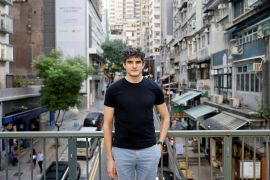 Samuel Bickett poses for a photo in black t-shirt and grey trousers on a bridge above a busy Hong Kong street lined with high-rise buildings