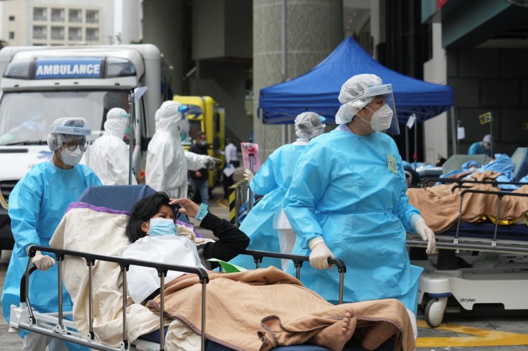 Medical workers in blue protective gear, hair nets and masks move a patient covered in a brown blanket on a trolley in a busy outdoor area of a Hong Kong hospital.
