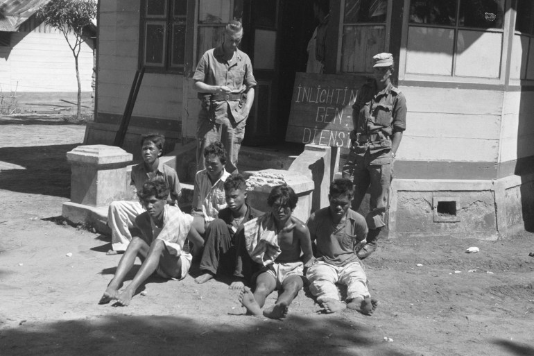 Two Dutch soldiers stand on the veranda of a house watching over a group of six Indonesian independence fighters who they plan to interrogate in 1946
