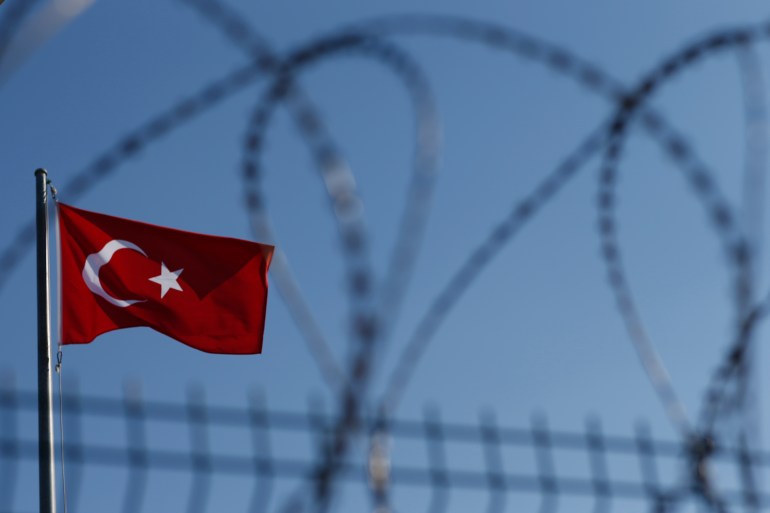 A Turkish flag fies in blue skies near a razor wire topped border fence