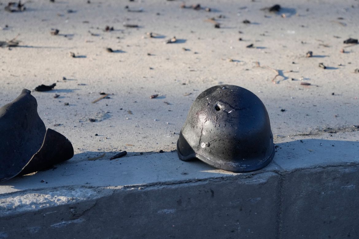 A soldier's helmet with a bullet hole near debris of burning military trucks
